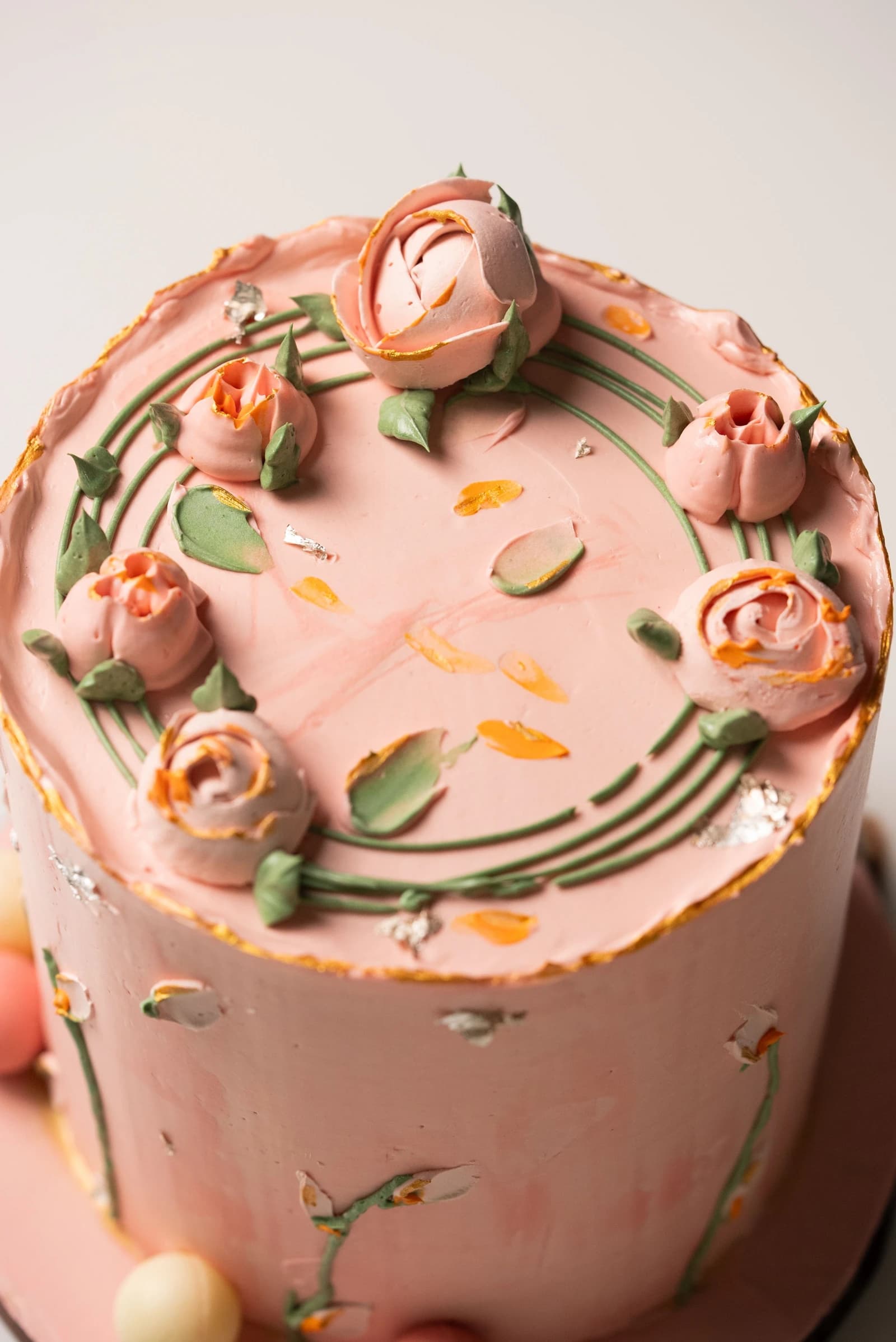 Smooth pink round cake with pink buttercream roses around the top, green stems and leaves, and small gold-colored accents.