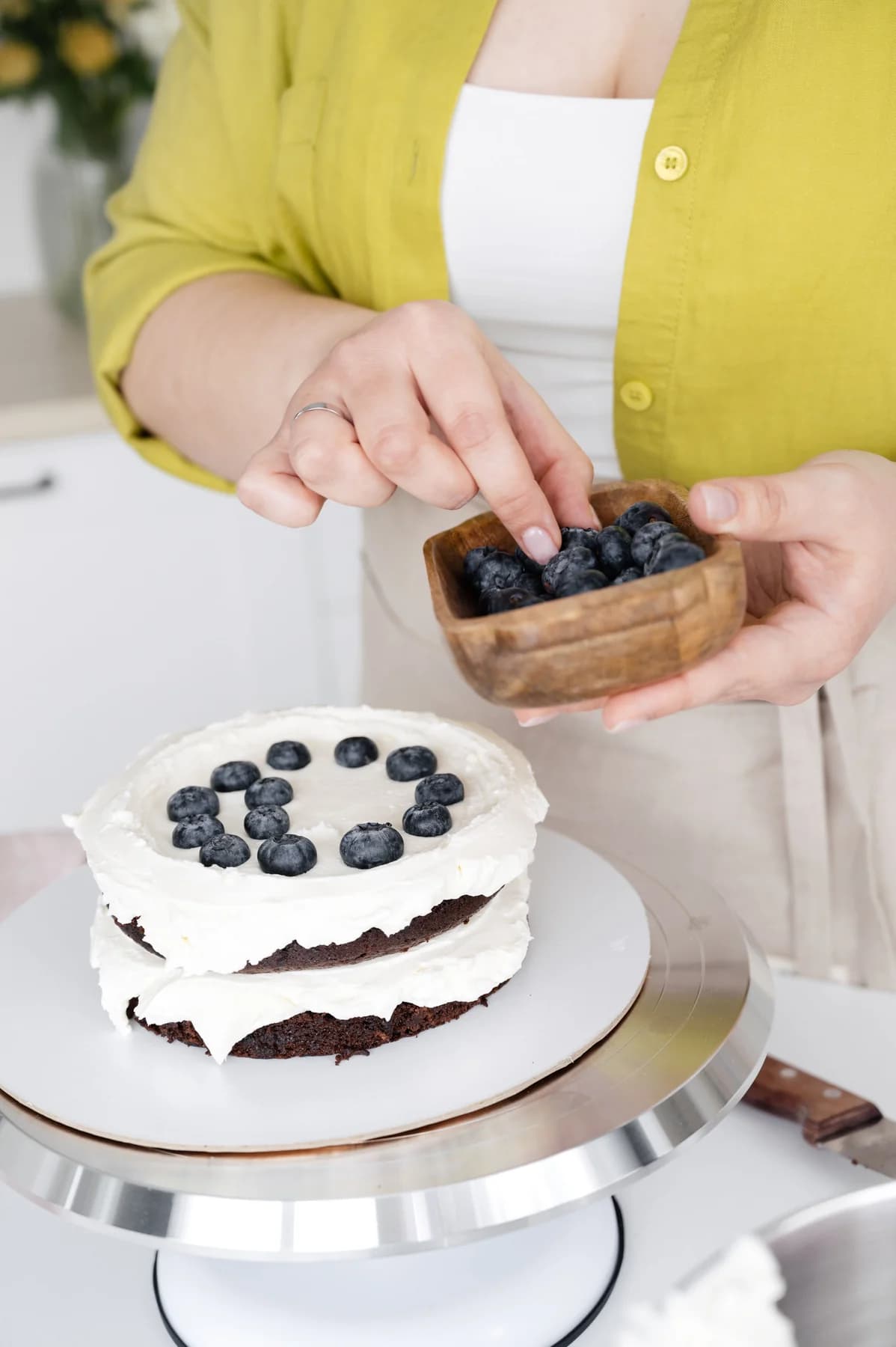 A baker arranges fresh blueberries on a cream-covered layer cake