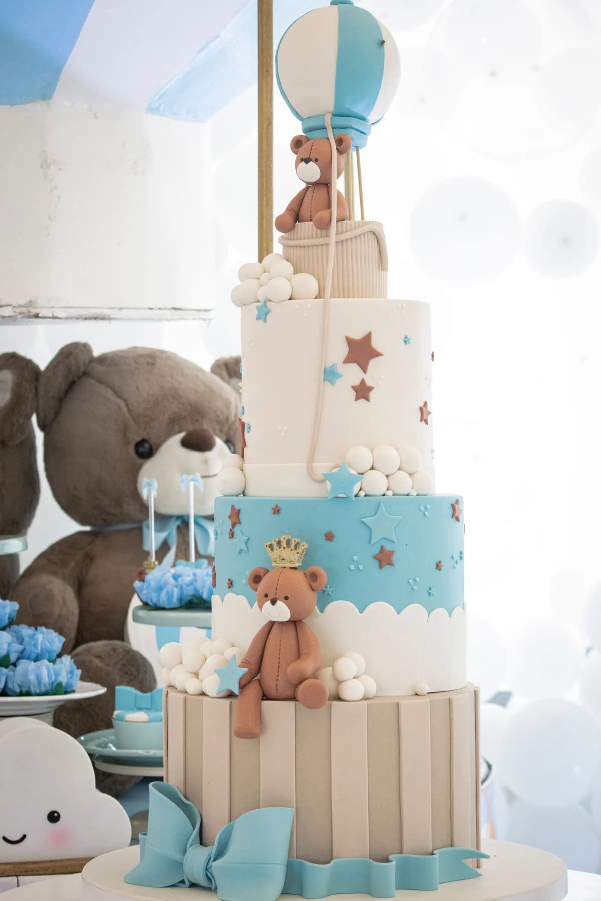 Tiered blue-and-white celebration cake with a teddy bear and balloon decorations