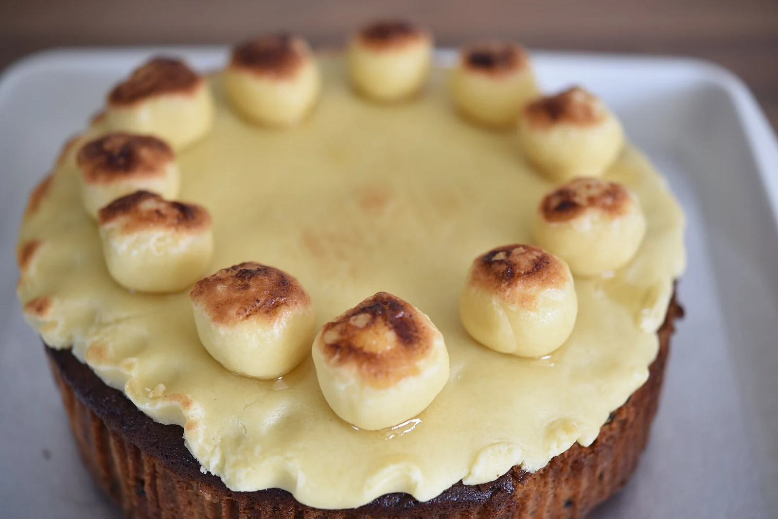A round dark brown cake on a white platter, covered with pale icing and topped with toasted-looking marzipan rounds arranged around the edge and across the top.