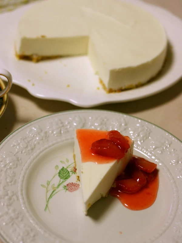 A triangular slice of pale cheesecake topped with glossy red fruit and sauce sits on a decorated plate; the remaining round cheesecake is on a scalloped platter behind it.