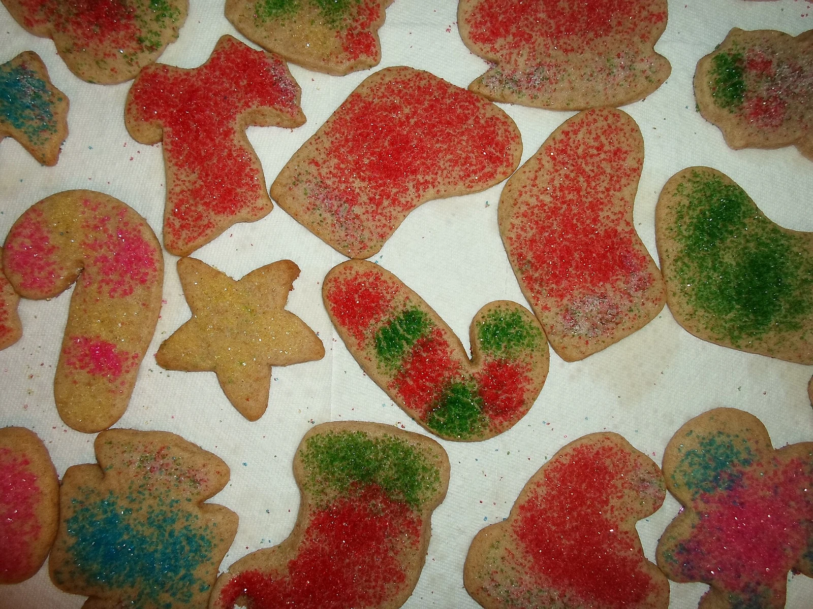 Assorted golden cutout cookies, including candy cane and mitten-like shapes, scattered with patchy red, green, pink, and blue glittering sugar on a white paper towel.