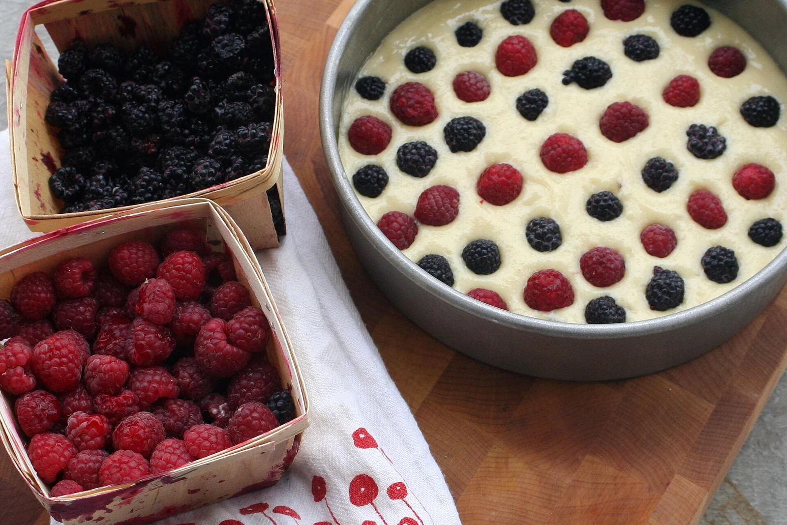 A round metal pan filled with pale cake batter and topped with scattered raspberries and blackberries; baskets of raspberries and blackberries sit beside the pan.