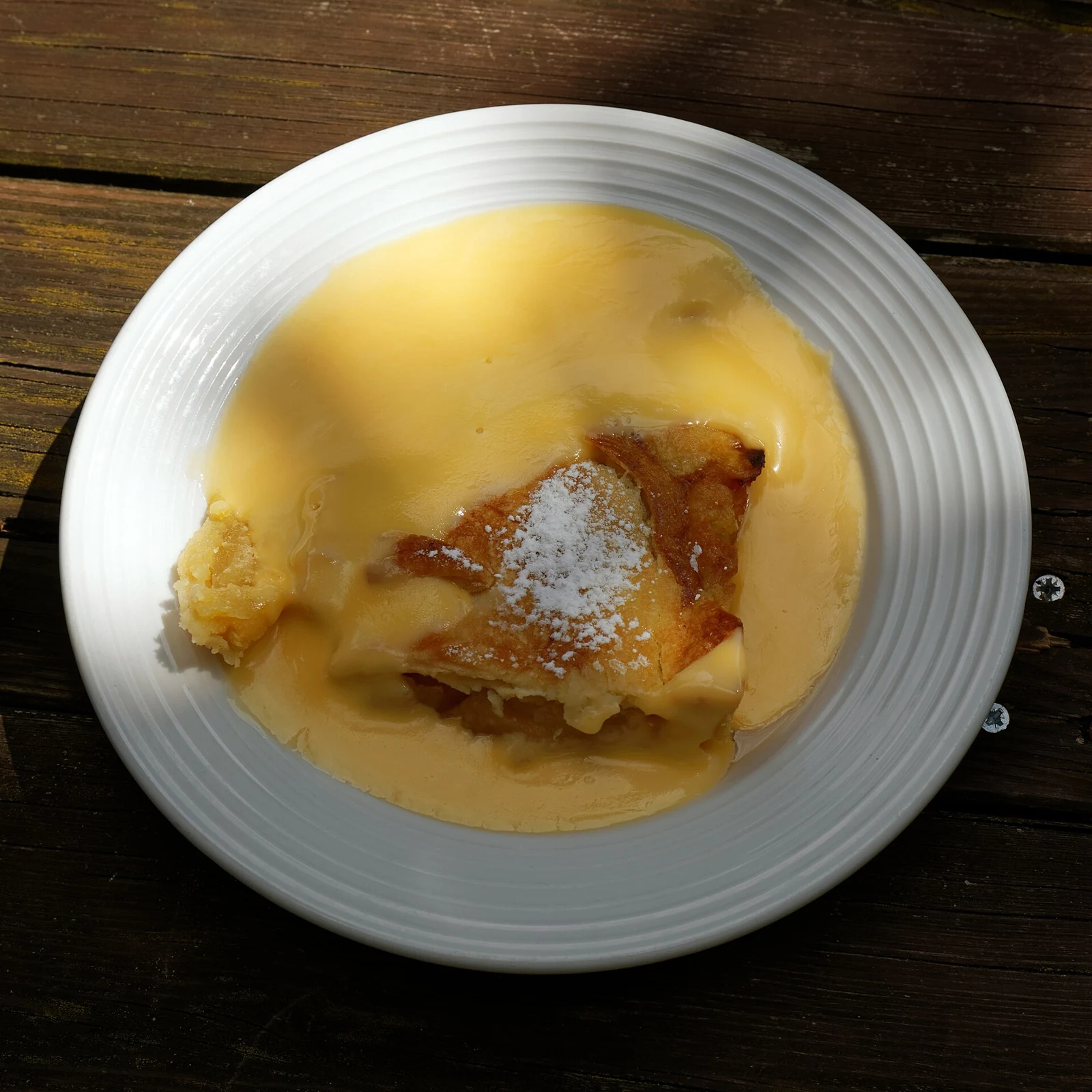 An irregular golden piece of baked tart dusted with powdered sugar sits in smooth yellow custard on a white plate, photographed on a dark wooden table.