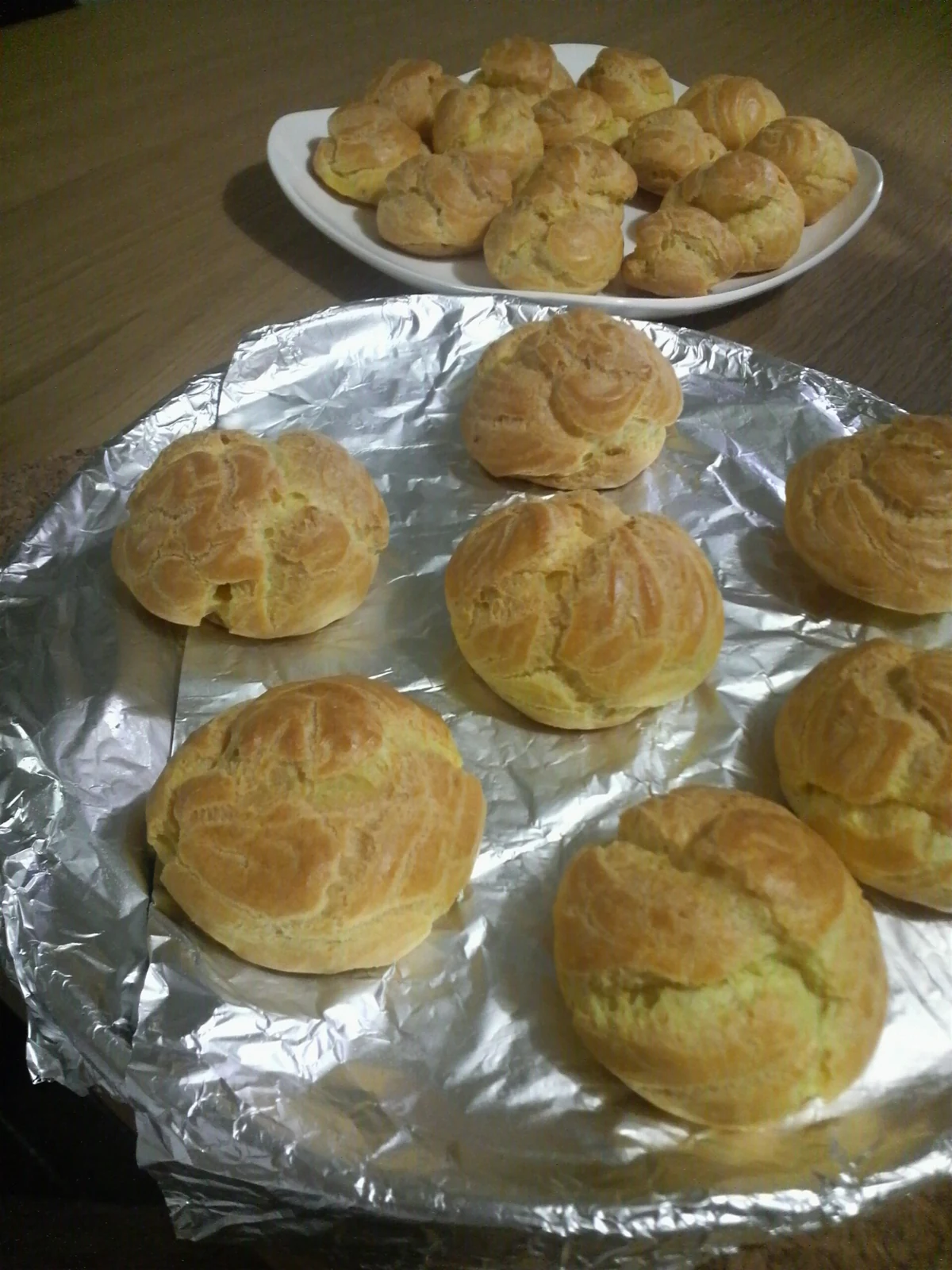Golden-brown round choux buns with naturally cracked tops rest on a foil-lined tray, with more small puffs on a white plate behind them.