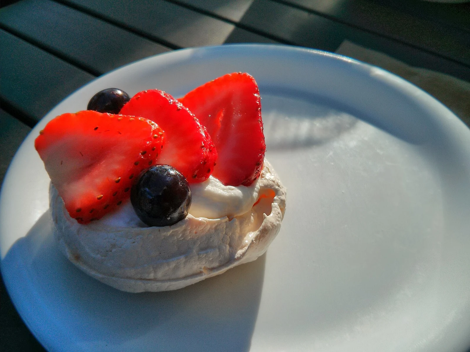 A small round pavlova with an irregular pale meringue shell, topped with white cream, sliced strawberries, and dark berries, on a light blue plate in direct sunlight.
