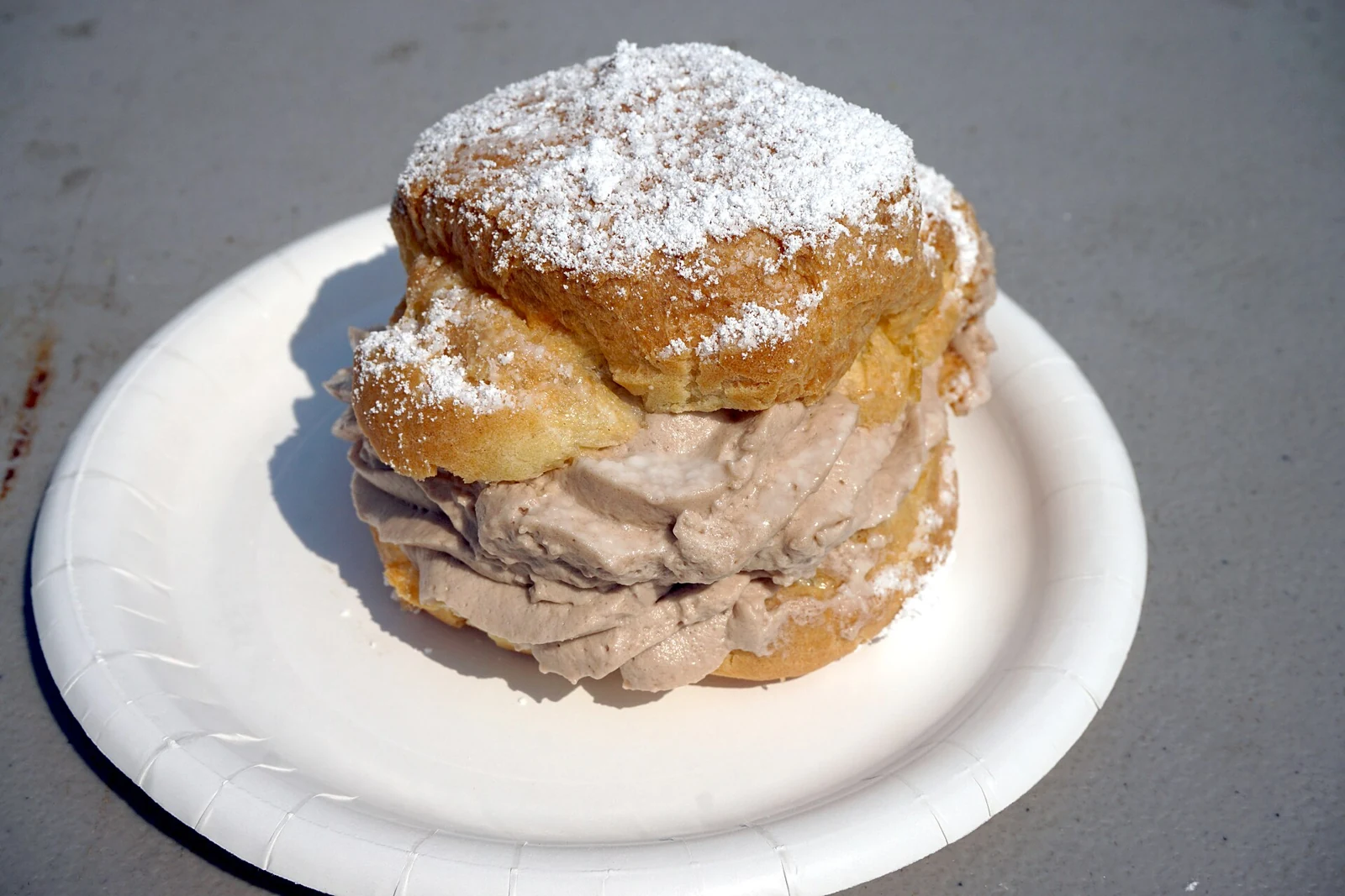 A large round cream puff with a domed, powdered-sugar-dusted choux top over a wider golden pastry base; pale cream shows between the pastry layers on a white plate.