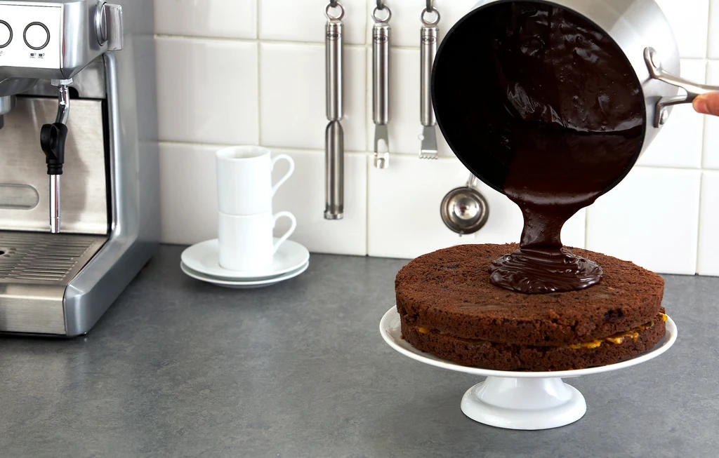 A two-layer round chocolate cake with a thin yellow filling visible between the sponges sits on a white pedestal stand as glossy dark ganache is poured from a pan onto the top.