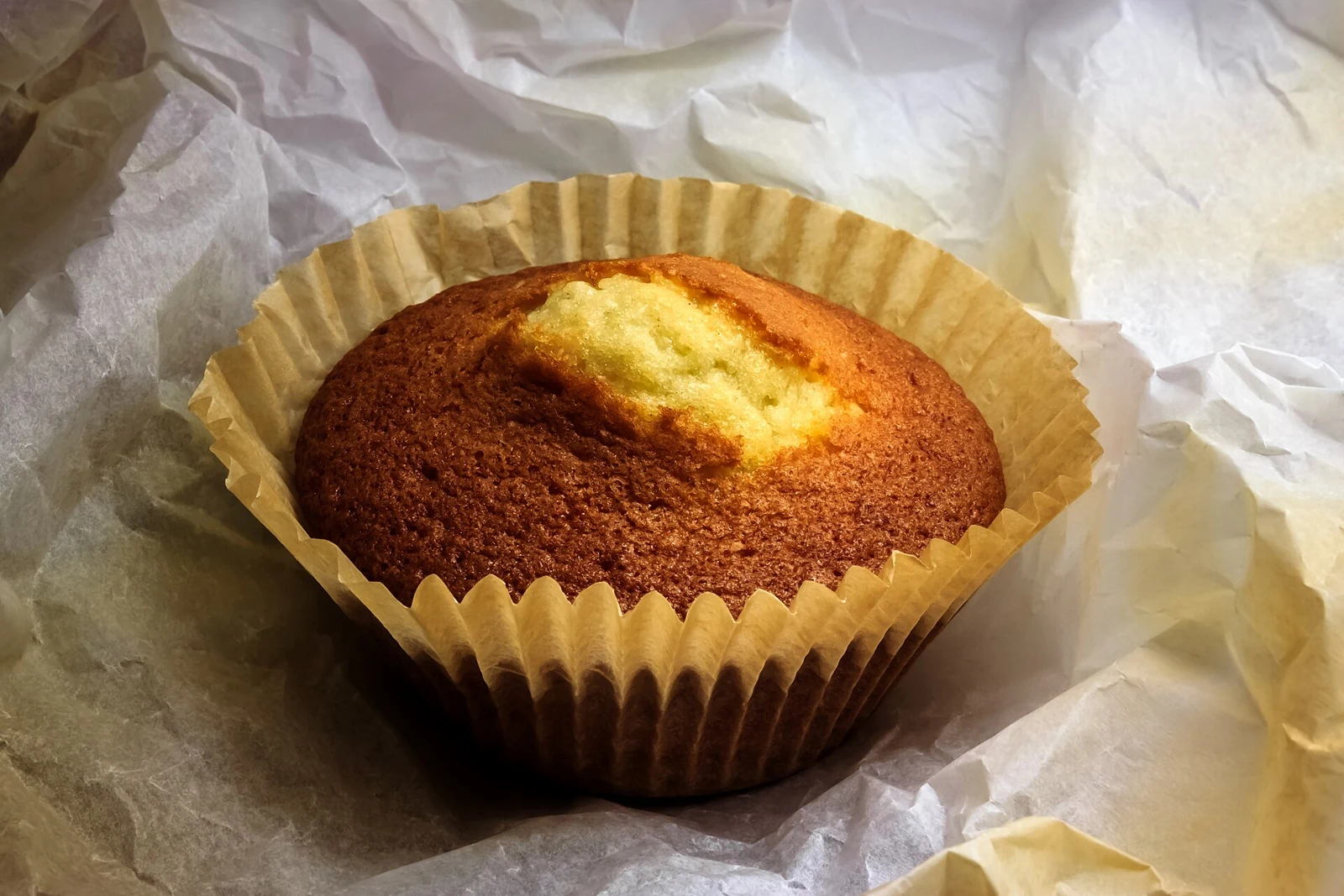 A single domed cupcake with a golden-brown top and a pale cracked patch at the center, resting in a fluted paper liner on crumpled baking paper.