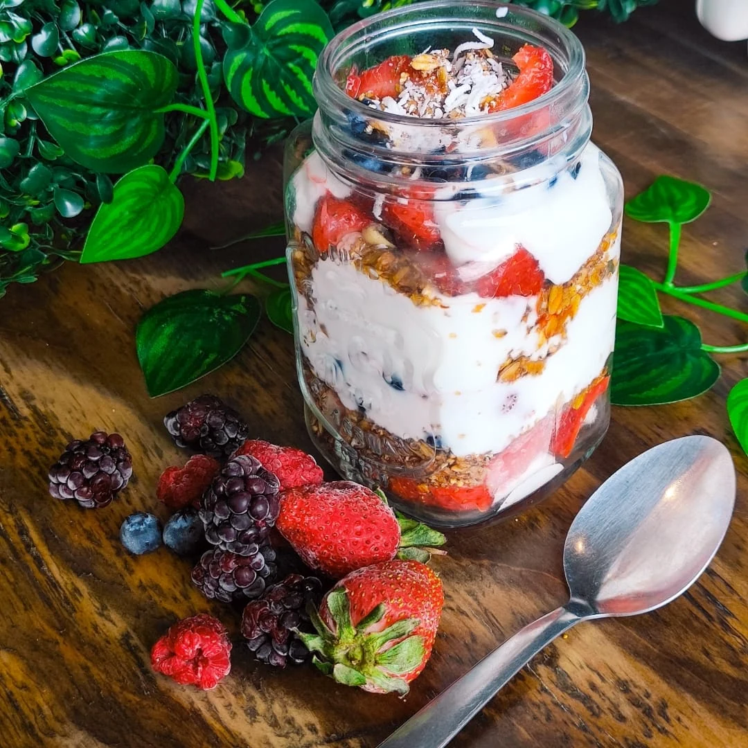 A clear jar of layered white cream, granola, and mixed berries topped with shredded coconut and fruit, with fresh berries and a spoon on a wooden table.