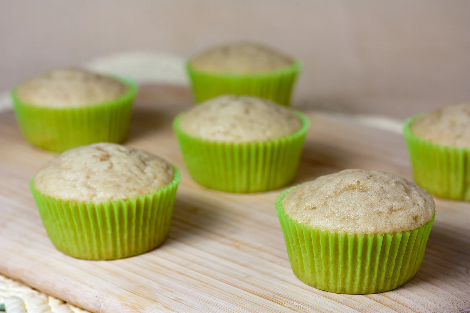 Several pale, gently domed cupcakes in bright green liners sit on a light wooden board; they are plain and unfrosted.