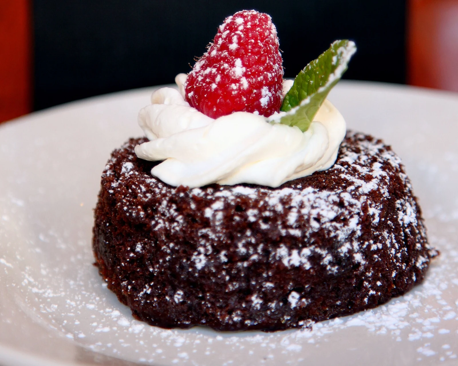 A round individual chocolate cake dusted with powdered sugar, topped with piped white cream, a raspberry, and a mint leaf, on a white plate.