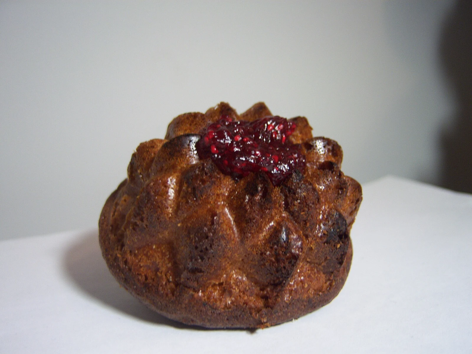 A small, browned fluted cake sits on a white plate, topped with glossy dark red berry preserve. The cake is intact, so its interior is not visible.