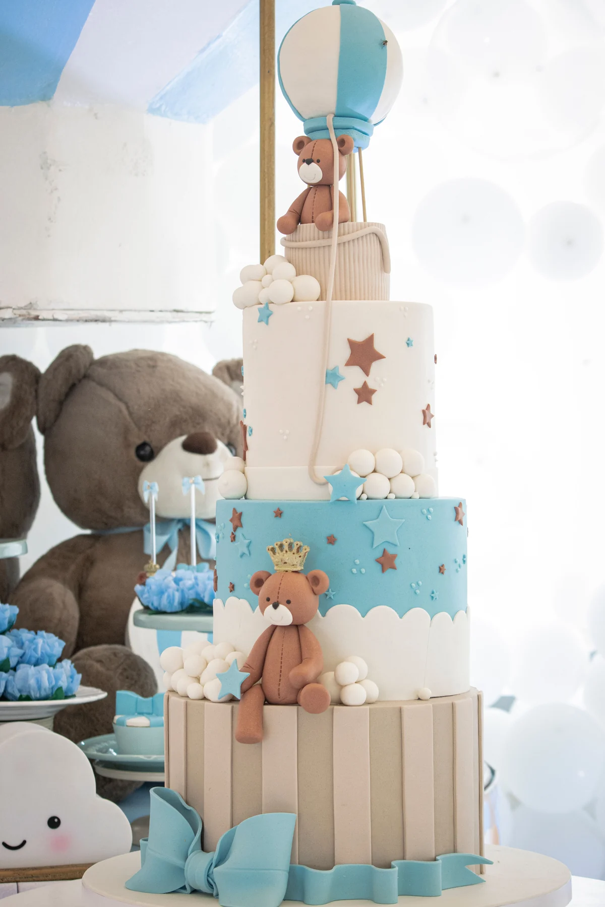 Tiered blue-and-white celebration cake with a teddy bear and balloon decorations