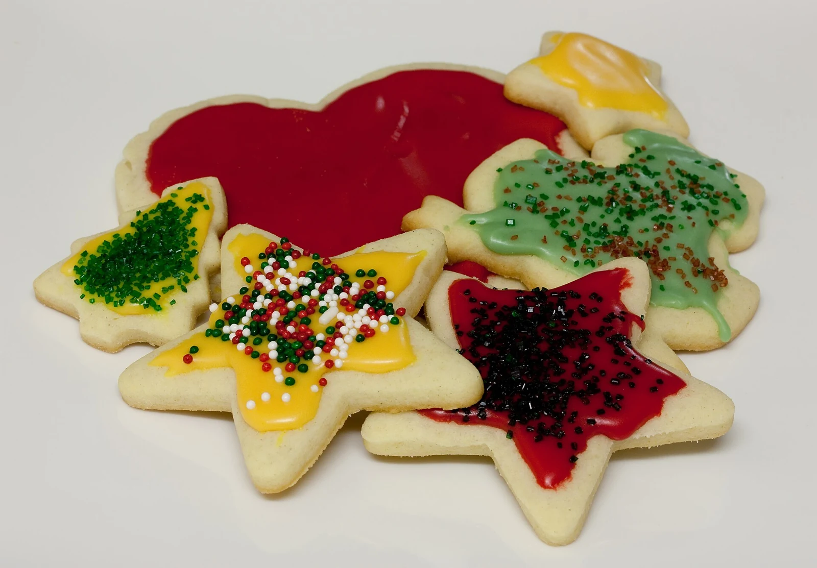 A pile of pale star-shaped cutout cookies decorated with smooth red, yellow, and green icing, colored sugar, and round sprinkles on a light surface.