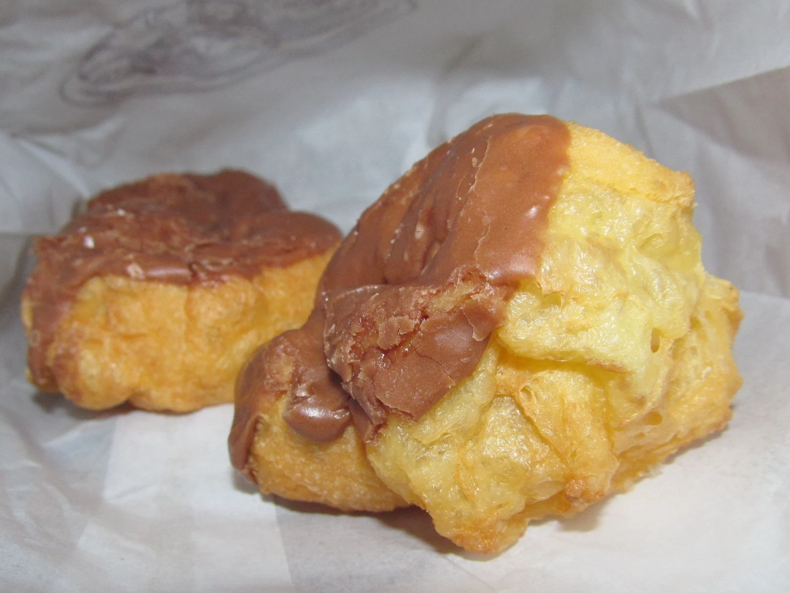 Two irregular, golden cream puffs on white paper, topped with glossy brown chocolate icing; no filling is visible.