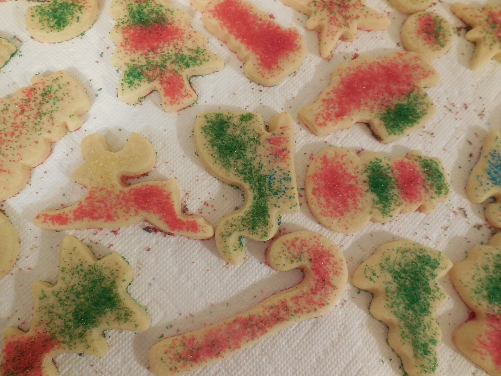 Assorted pale, flat cut-out cookies in animal and simple shapes, scattered with red and green colored sugar on a white paper towel.