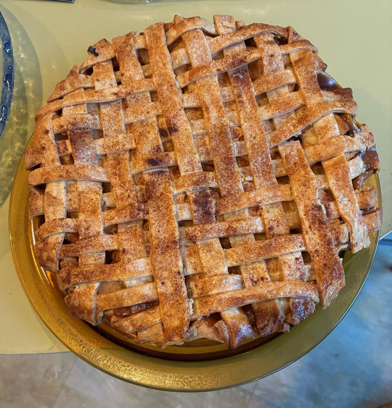 A round apple pie on a gold-colored platter, covered with a wide woven pastry lattice dusted with brown spice; glossy filling shows through in places.