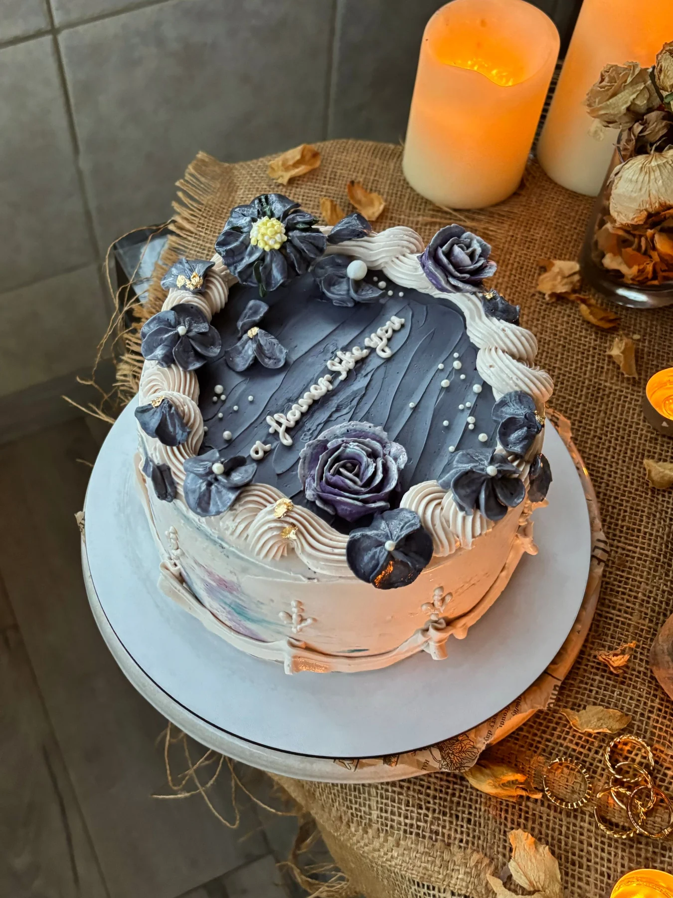 Dark floral cake with sculpted roses and white piped borders, beside candles