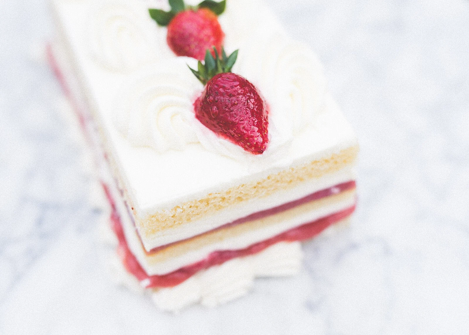 A layered strawberry cake slice with pale sponge, pink filling, white cream and a whole strawberry on top, photographed on a light surface.