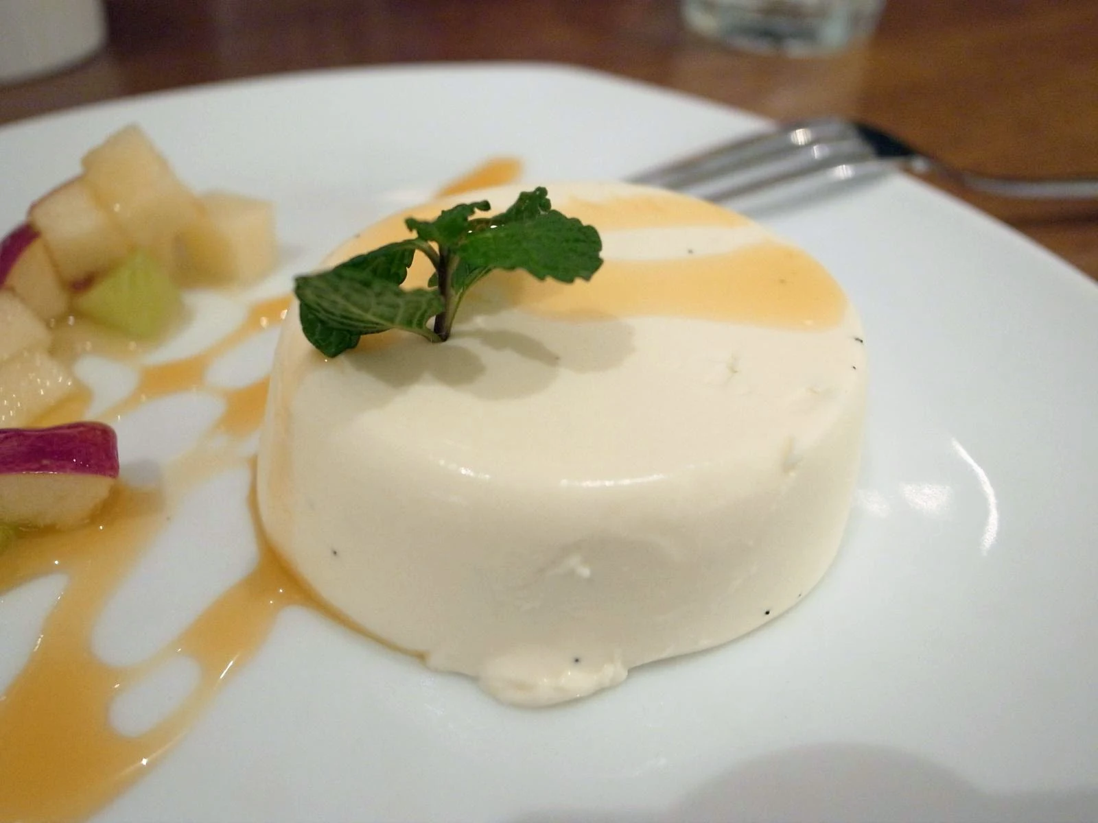 A smooth, pale panna cotta with amber sauce and a mint sprig, plated beside diced fruit on a white plate.