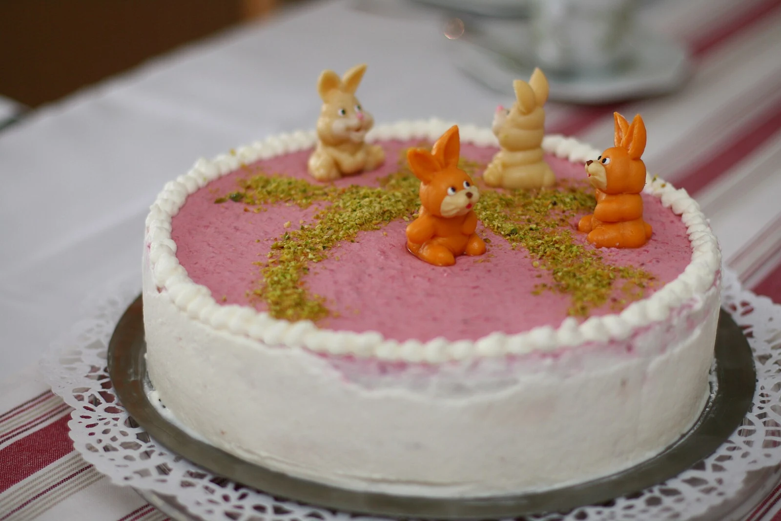 A round, single-tier cake with smooth white frosting on the sides, a pink top, a piped white border, a curved trail of chopped green nuts, and small rabbit-shaped decorations.