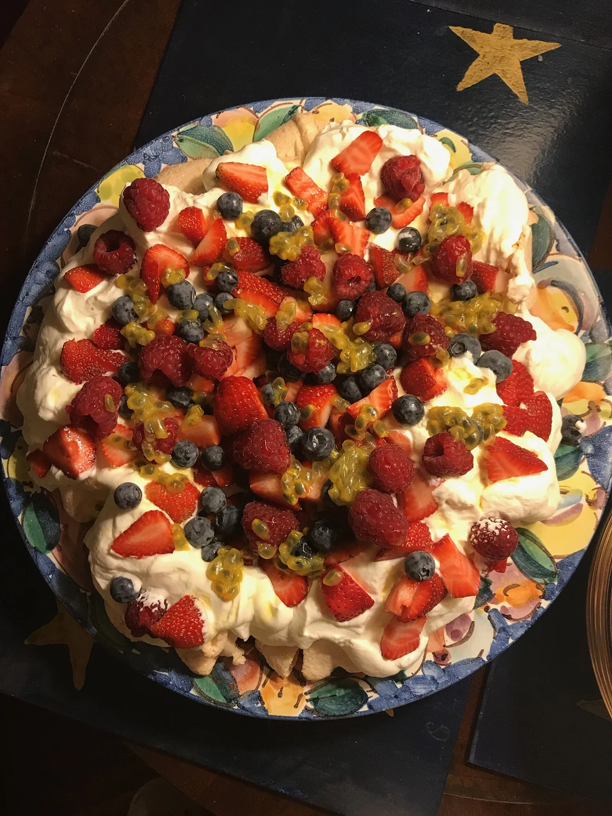 A large round pavlova-style dessert on a colorful floral plate, topped with white cream, sliced strawberries, raspberries, blueberries, and golden passion fruit pulp.