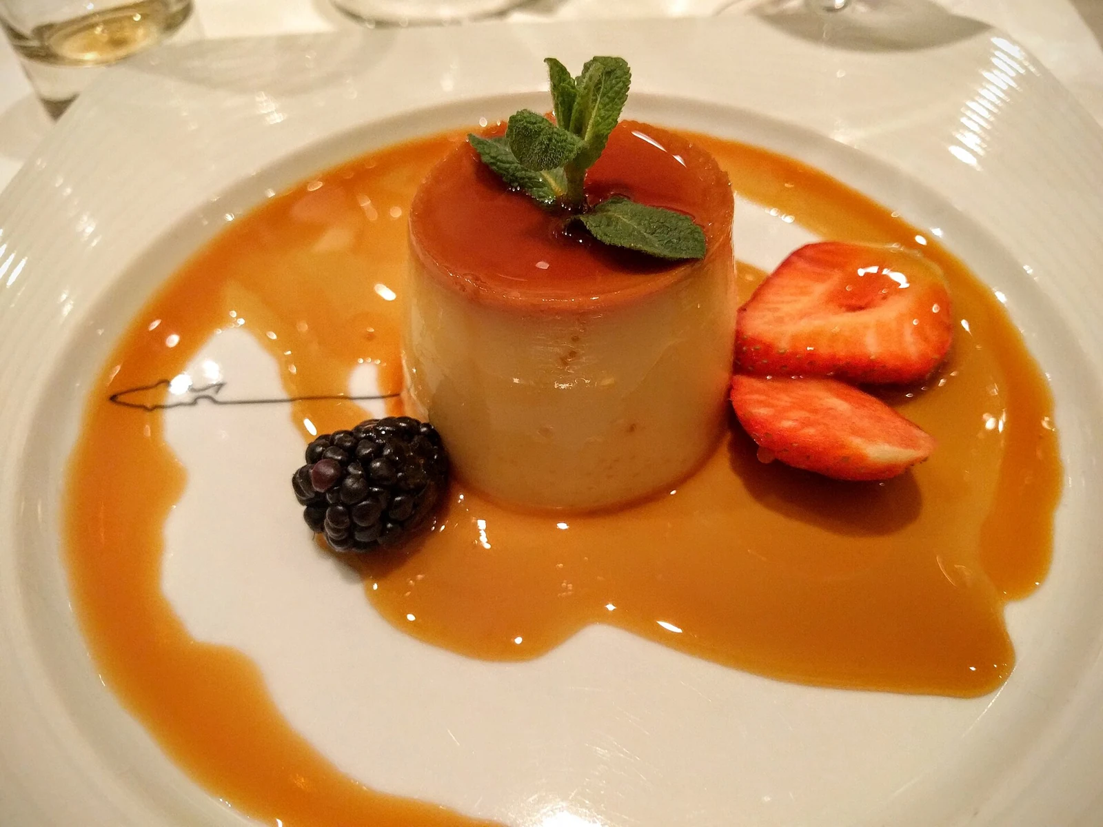 A pale cylindrical panna cotta topped with glossy amber sauce and mint, plated in a pool of sauce with sliced strawberries and a blackberry.