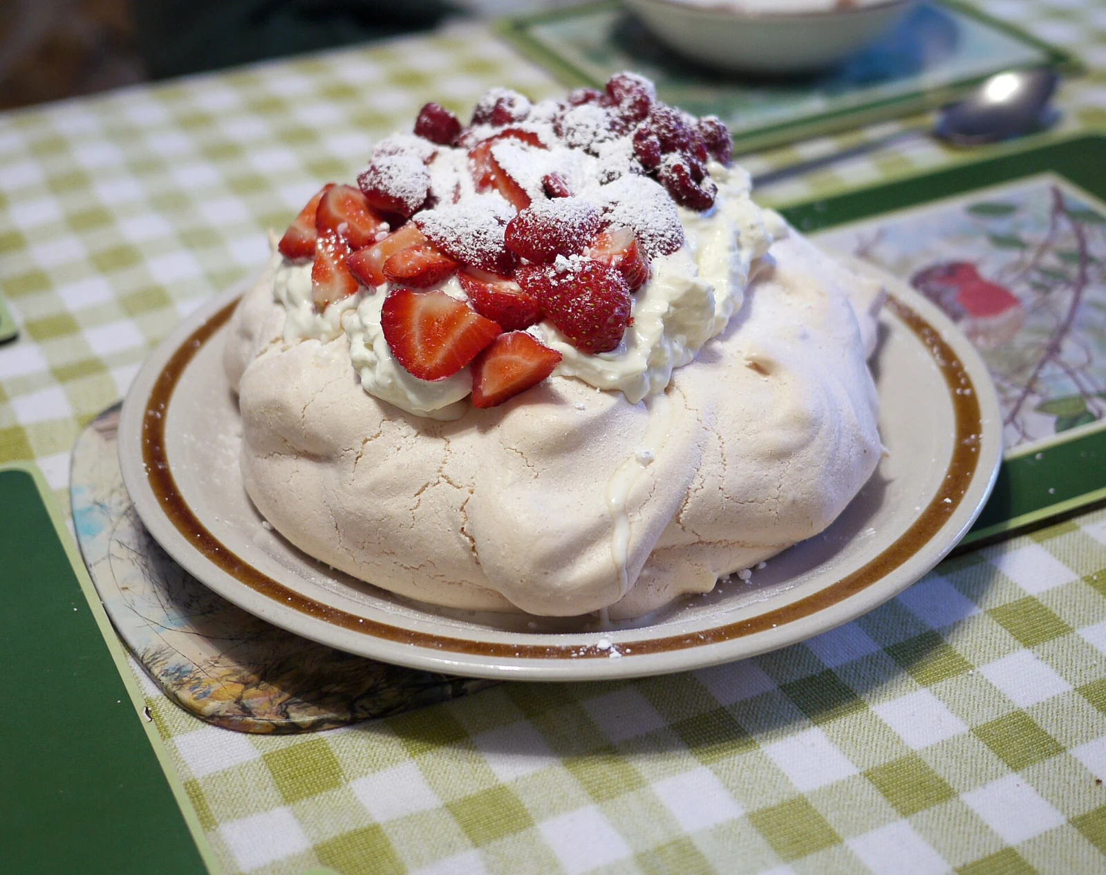 A round, pale meringue pavlova on a plate, topped with white cream, sliced strawberries and other red berries, then dusted with powdered sugar.
