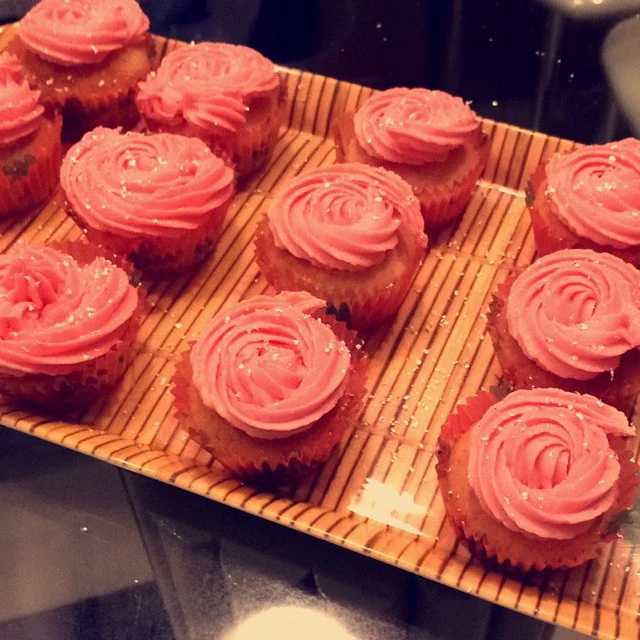 Vanilla cupcakes in paper liners arranged on a bamboo mat, topped with coral-pink piped rosette frosting and fine sugar sprinkles.