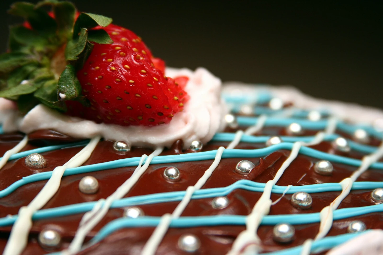 Close-up of a rectangular chocolate-glazed cake decorated with crossing blue and white icing lines, silver sugar beads, and a strawberry with green leaves on a pale cream rosette.