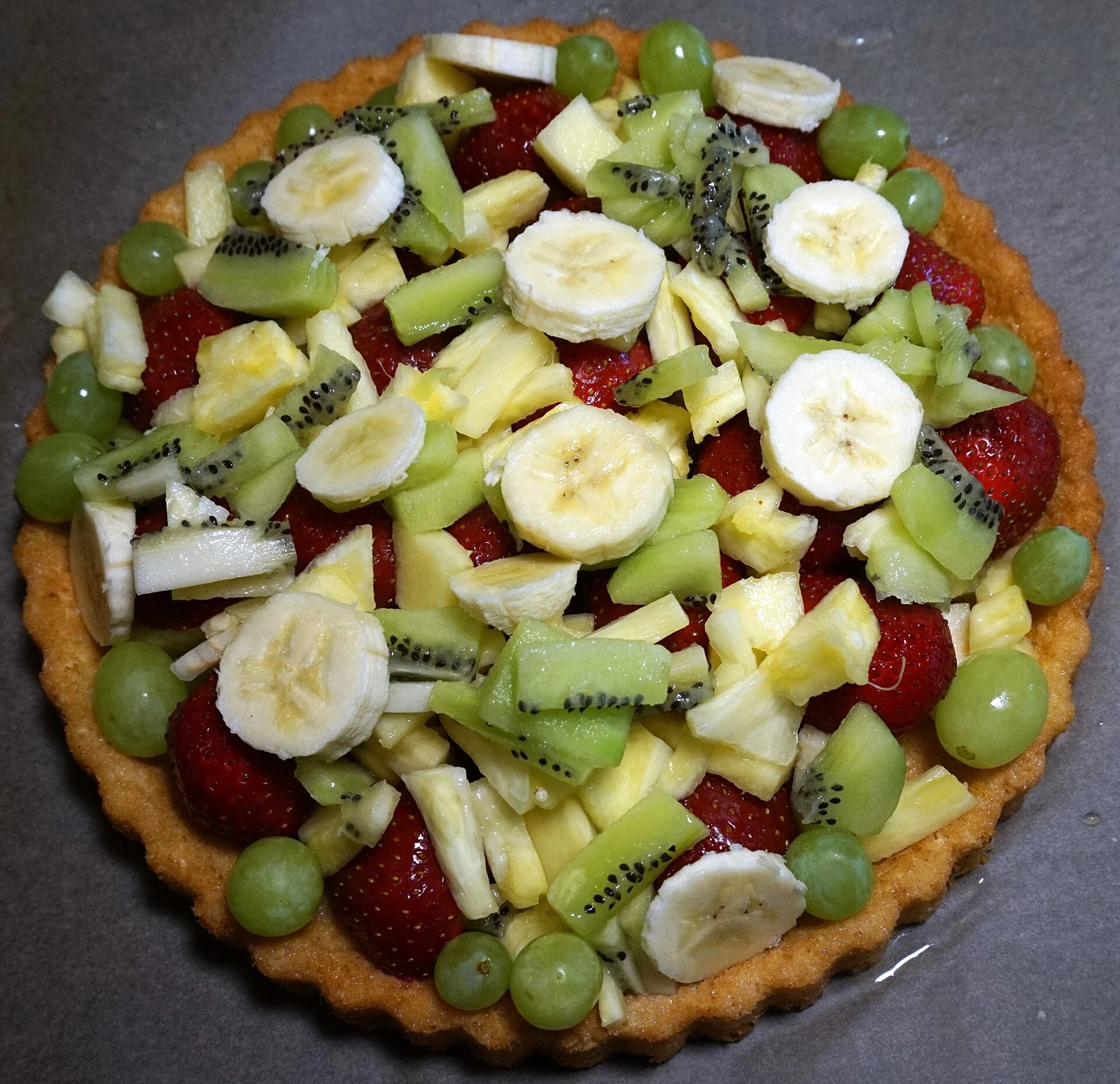 A round tart with a golden fluted crust and a generous topping of sliced strawberries, banana, kiwi, pineapple, and pear, with green grapes.