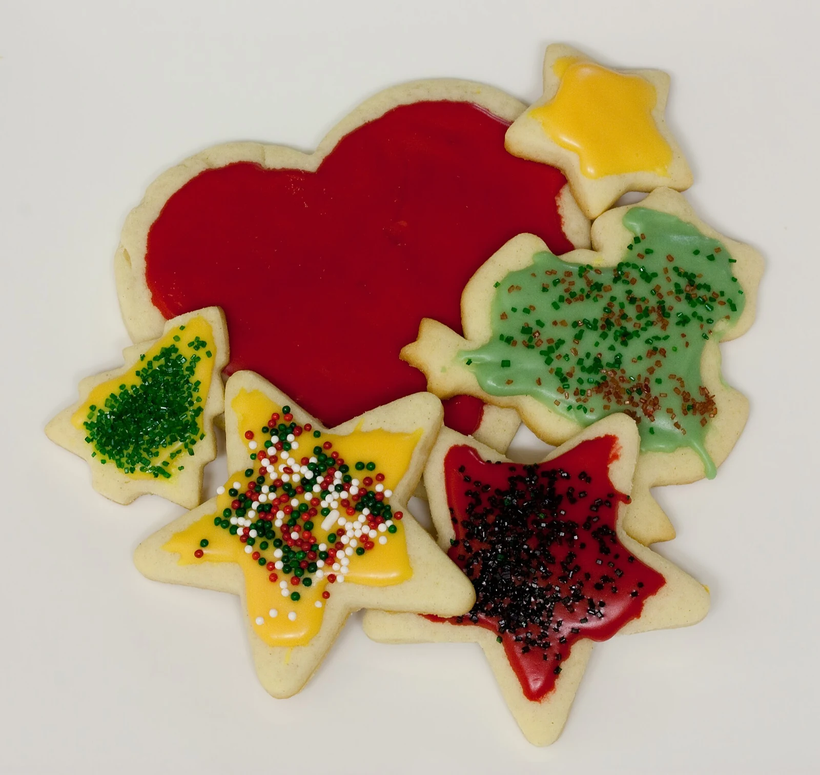 A pile of pale cut-out cookies on a light surface, including stars, trees, and a large rounded shape, decorated with red, yellow, and green icing, colored sugar, and small sprinkles.