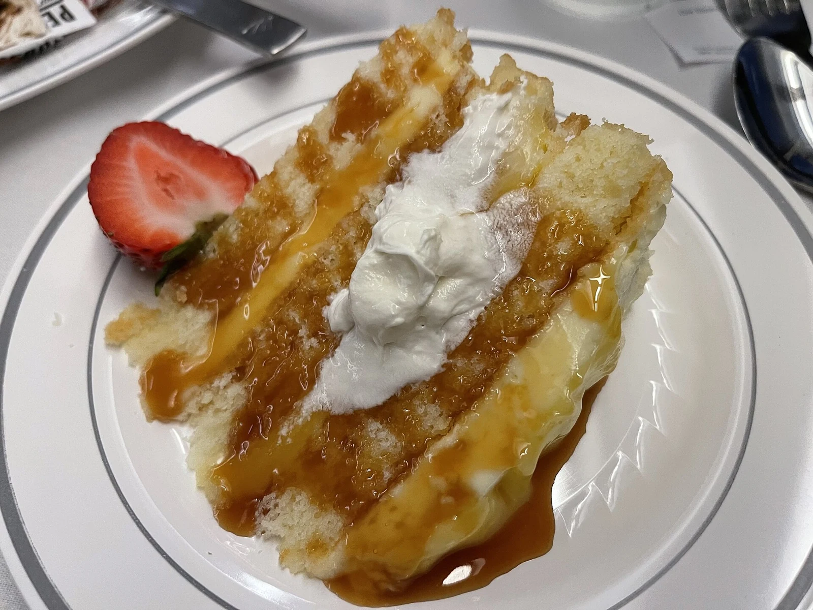 An irregular slice of pale layered sponge cake on a white plate, topped with a central dollop of white cream and glossy amber sauce, with a strawberry half beside it.