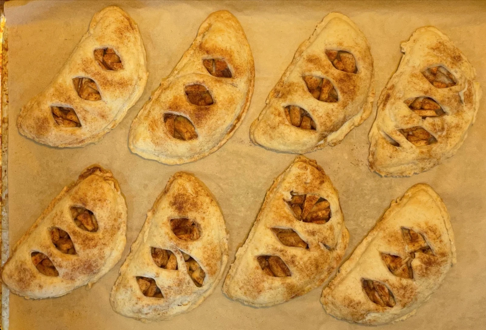 A tray of irregular oval hand pies with sealed edges and cut openings, showing apple pieces beneath lightly browned pastry dusted with cinnamon.