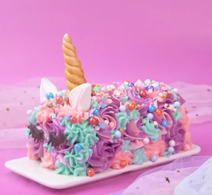 A rectangular cake covered with pink, purple, and mint piped frosting, scattered candy pearls, candy eyes, and a golden horn with pale ears, displayed on a white board against a pink background.