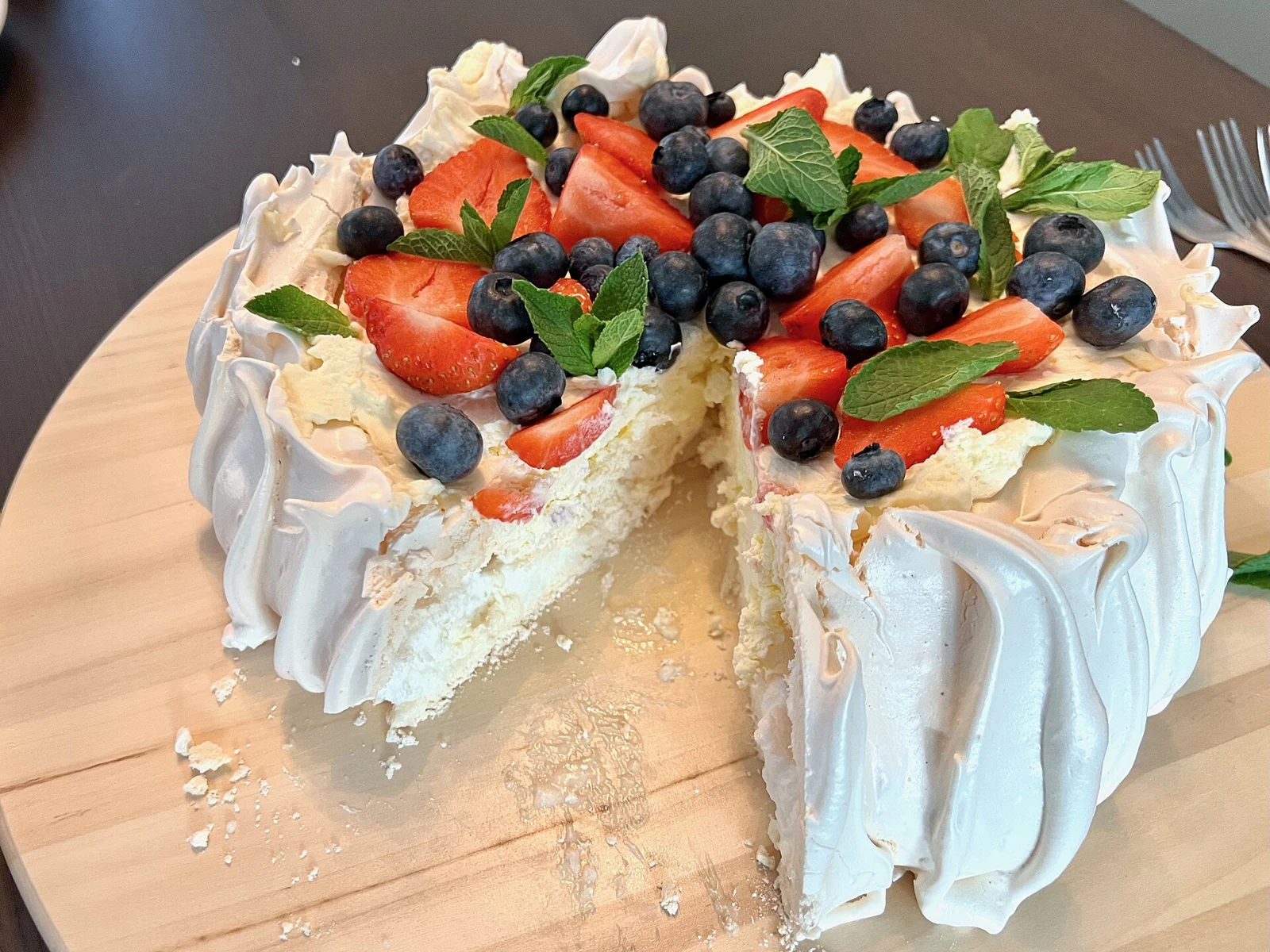 Round pavlova with a tall, ruffled white meringue shell, topped with whipped cream, strawberries, blueberries, and mint leaves. A wedge has been cut away, exposing the pale interior.