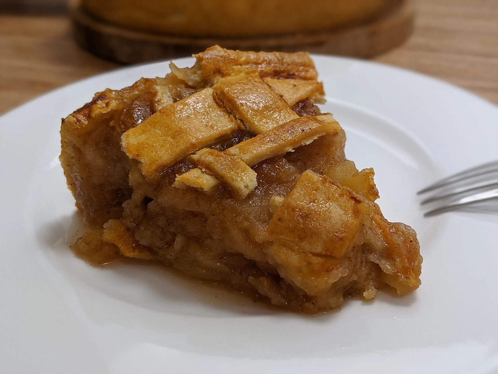 A thick triangular slice of apple pie with a golden, uneven pastry top and soft spiced apple filling sits on a white plate beside a fork.