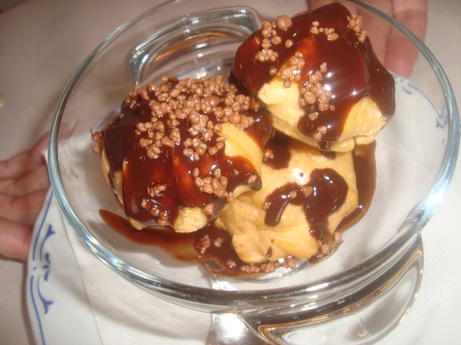 Several small, round golden cream puffs piled in a clear glass dish, topped with glossy dark chocolate sauce and small chocolate sprinkles.