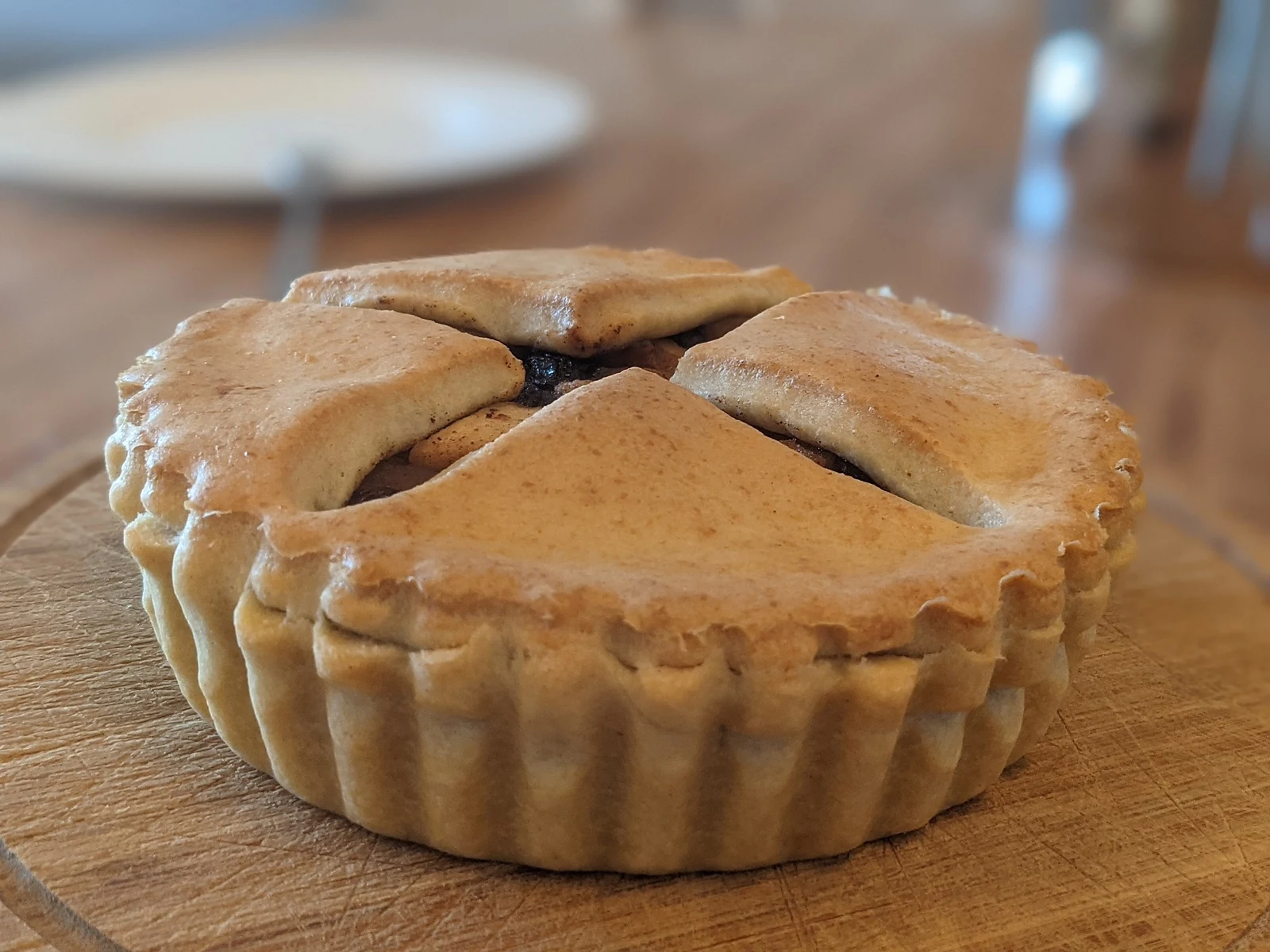 A round tart with fluted golden pastry sides and broad pastry pieces covering most of the top, with small gaps showing dark filling, on a wooden board.