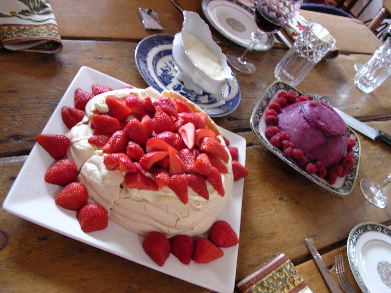 A large, irregular round pavlova topped with white cream and abundant strawberries sits on a white platter, with extra strawberries near its base. A bowl of purple berry dessert is beside it on a wooden table among serving dishes and glassware.