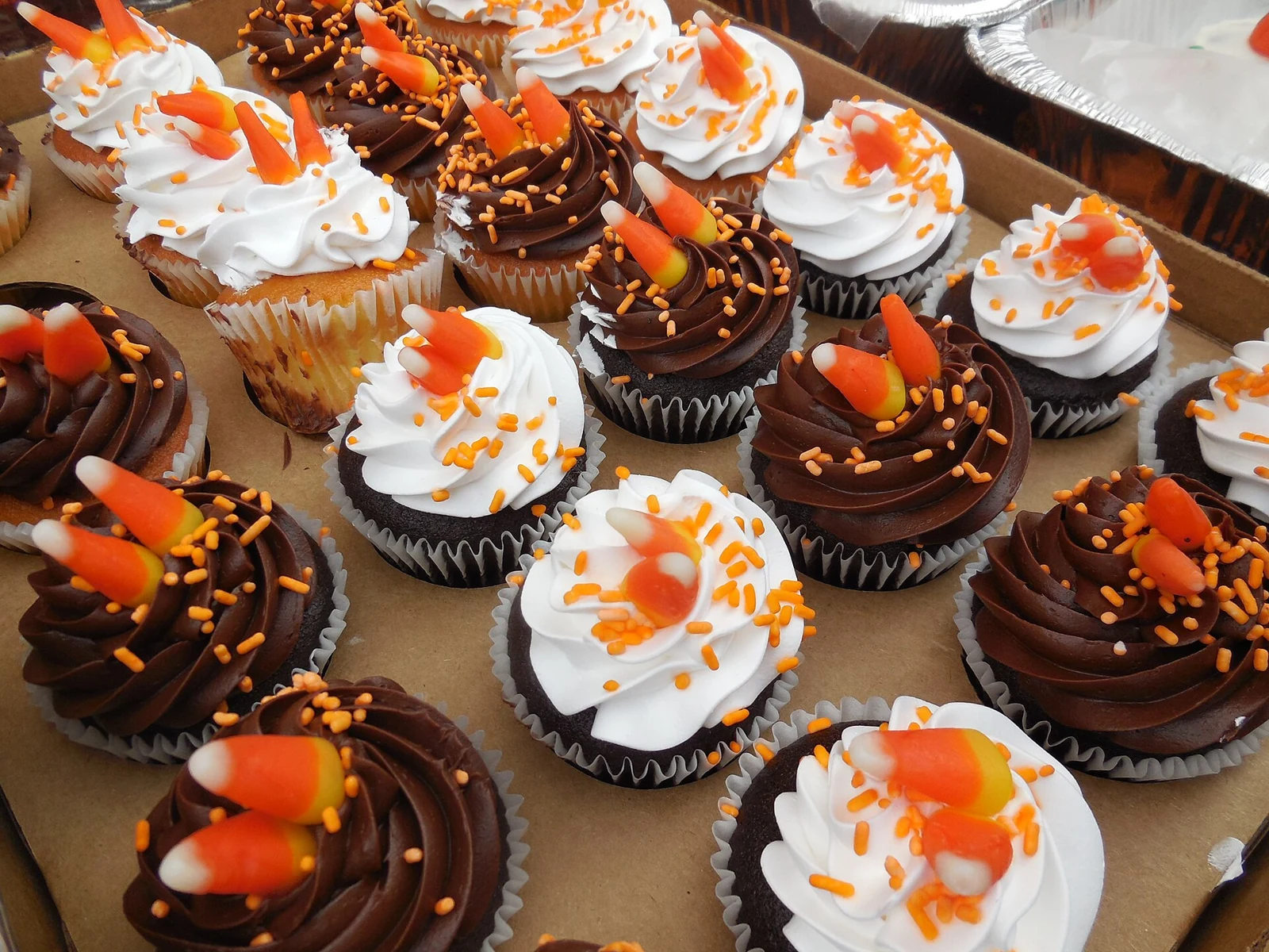 A tray of individual cupcakes in paper liners, topped with alternating white and chocolate piped frosting, orange sprinkles, and candy corn pieces.