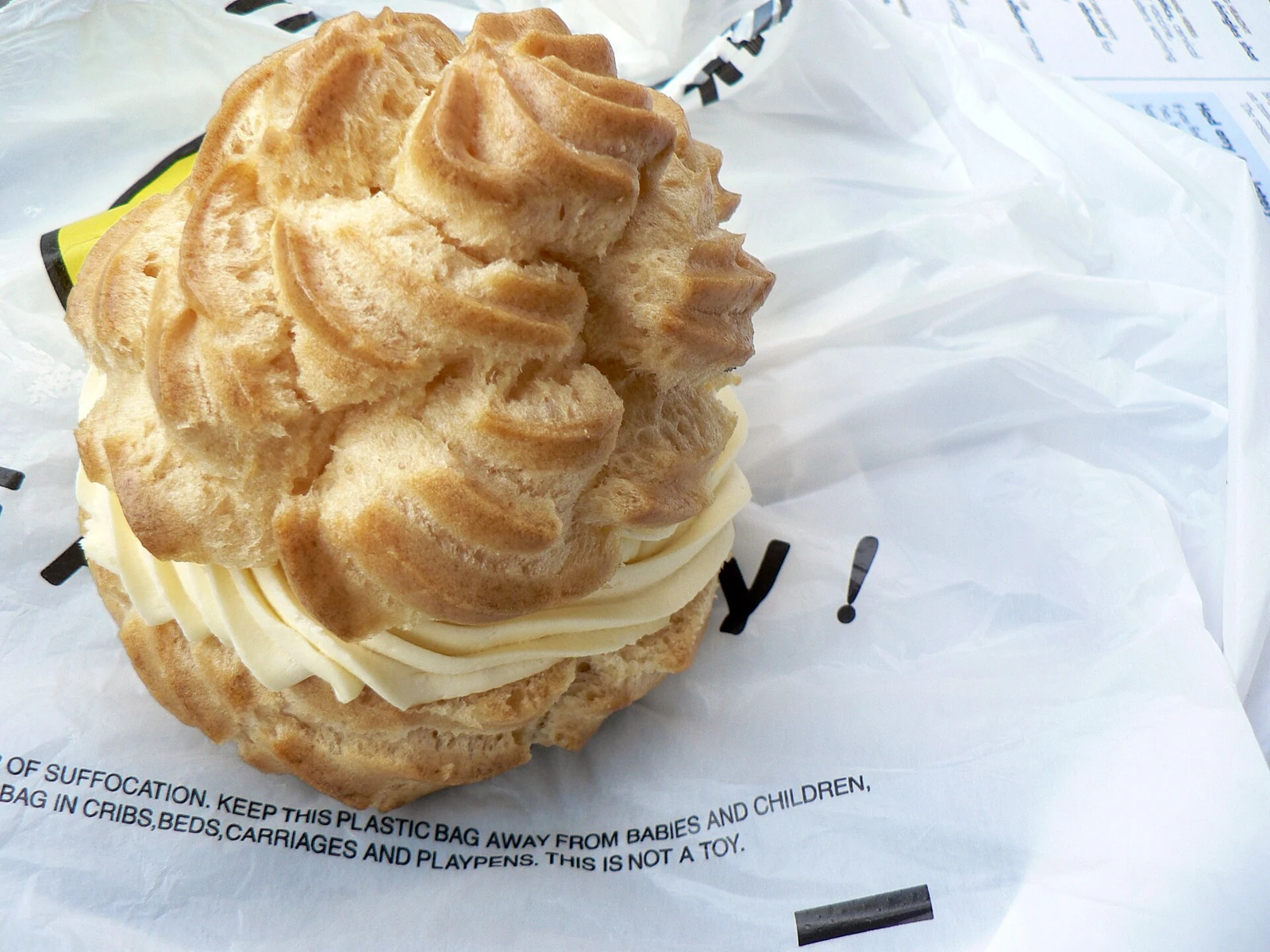 A large, irregularly stacked golden choux pastry with ridged piped pieces and a thick band of pale cream, resting on printed white packaging.