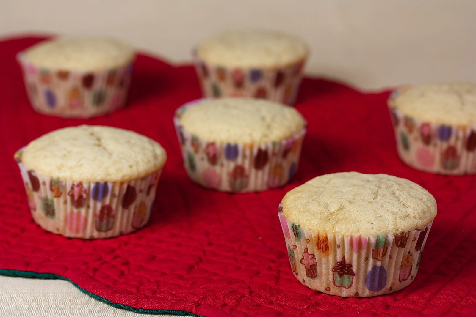 Several plain, pale-golden cupcakes in colorful patterned paper liners sit on a red cloth.