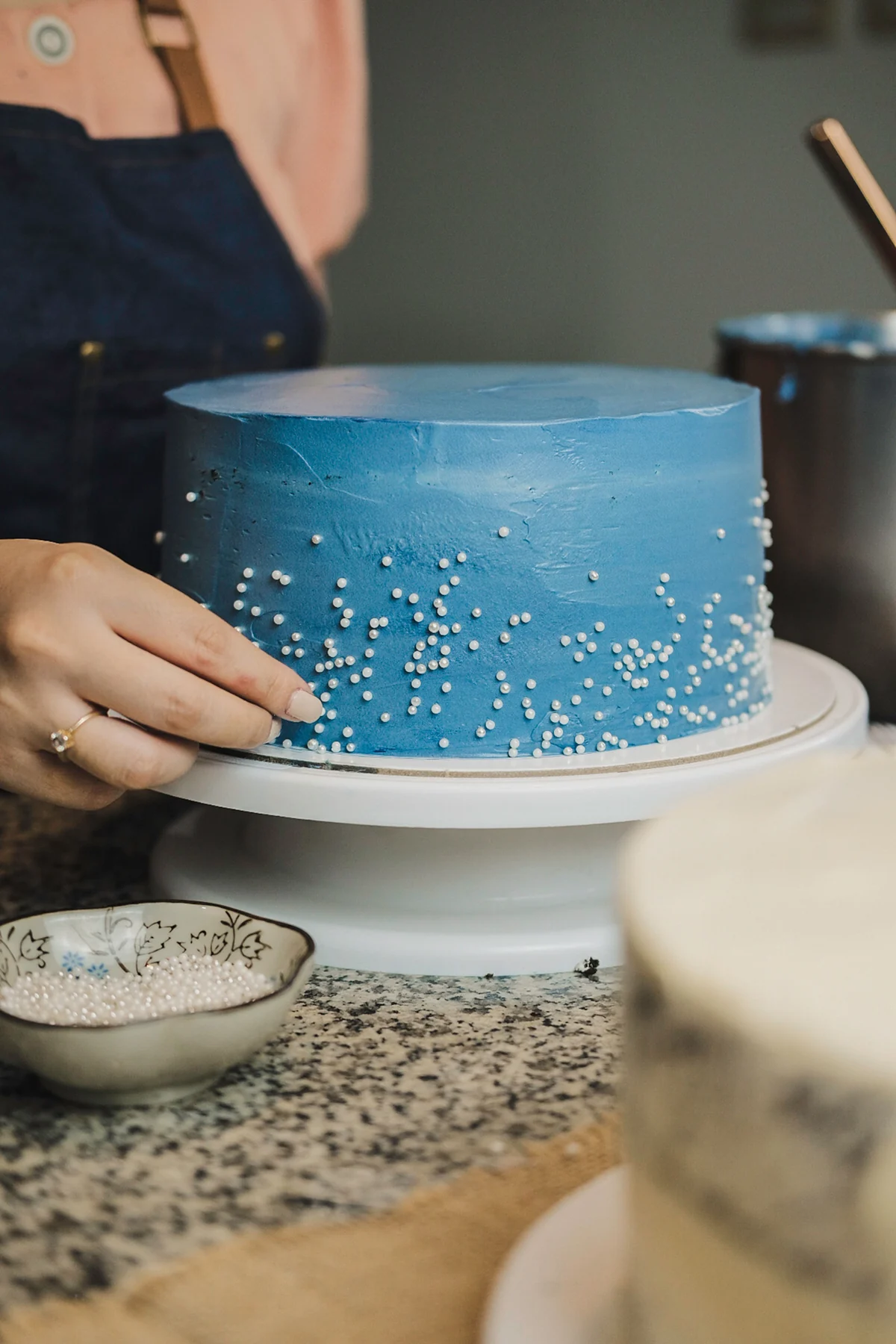A baker adds small white pearls to a blue-frosted cake
