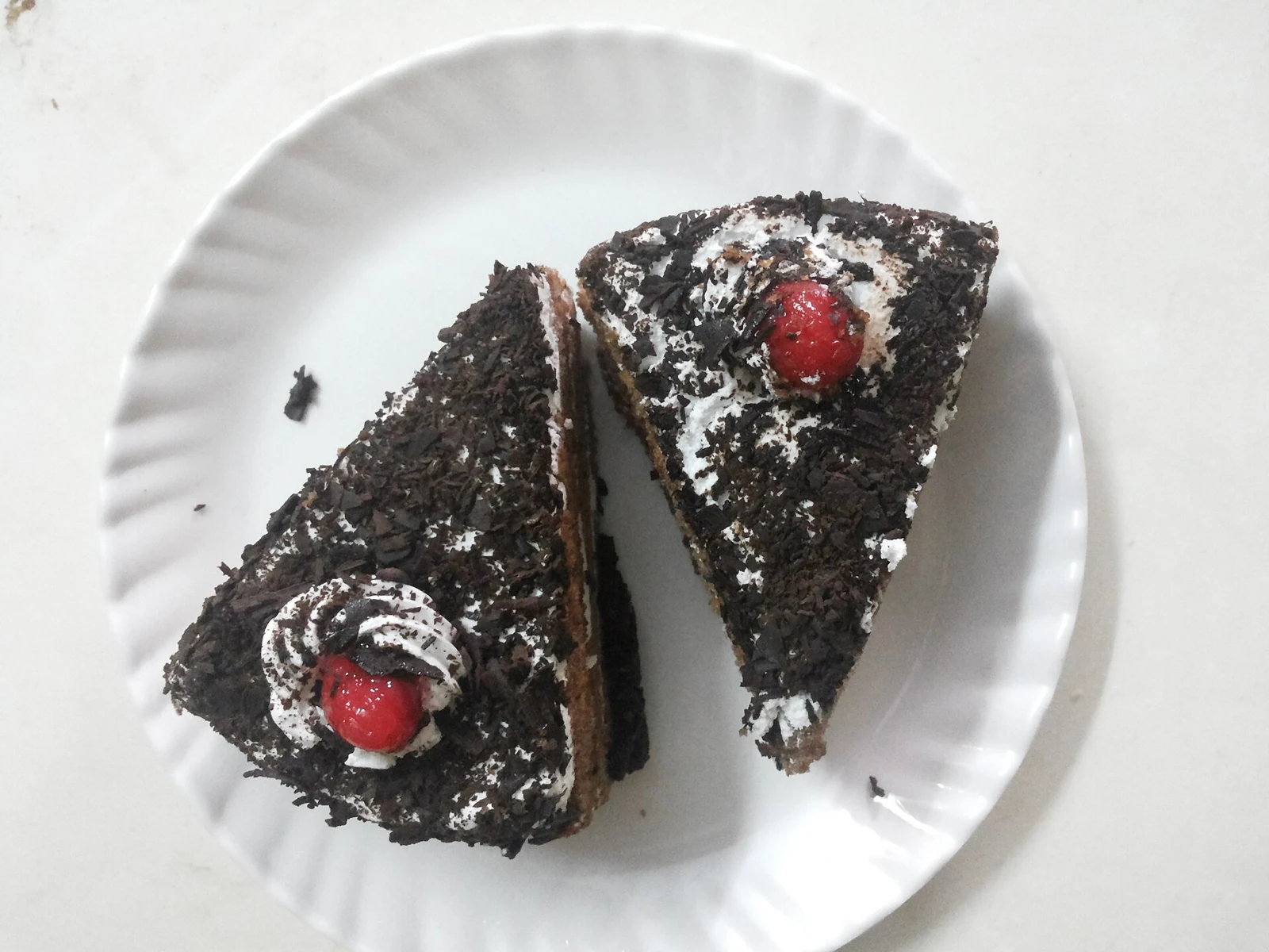 Two triangular slices of chocolate layer cake on a white plate, covered with rough dark chocolate shavings and decorated with white cream swirls and red cherries; chocolate sponge and pale filling show along the cut edges.